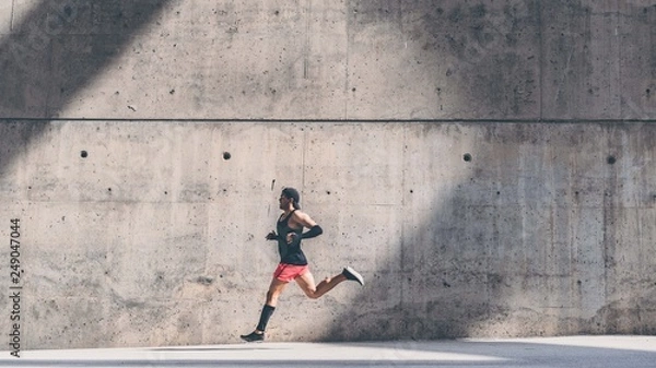 Fototapeta Muscular man athlete sprinter running fast,exercising outdoors,jogging outside against gray concret background with copy space area for text message or ad content.Side view,full length