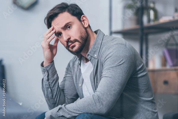 Fototapeta selective focus of handsome upset man sitting, touching face and grieving at home
