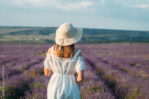 Fototapeta A young girl with a hat and a white dress stands with her back in a lavender field