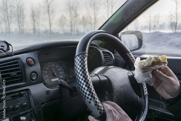 Fototapeta In the car, a man holds a bitten bun in his hand and drives the car. on the background of the steering hand with cheesecake.