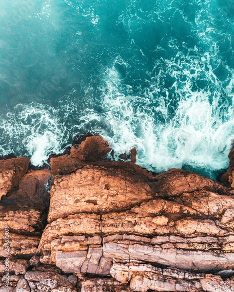 Fototapeta Turquoise blue cold sea waves hitting and crashing on warm colors rocks. Beautiful dramatic aerial drone shot abstract texture