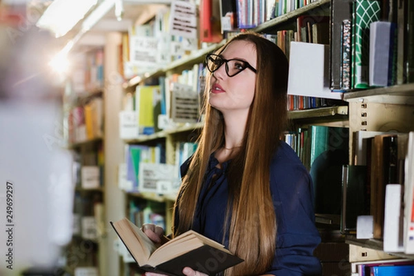 Fototapeta Beautiful young woman wearing glasses in the library among the bookshelves holding a book in her hands and reading, studying.