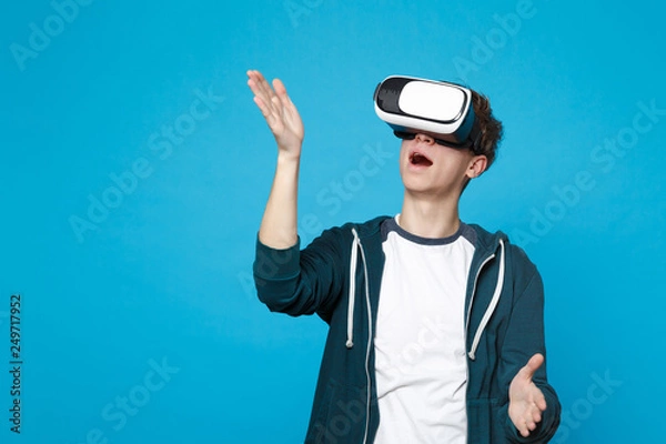 Obraz Amazed young man in headset touch something like push click on button, pointing at floating virtual screen isolated on blue background. People sincere emotions, lifestyle concept. Mock up copy space.