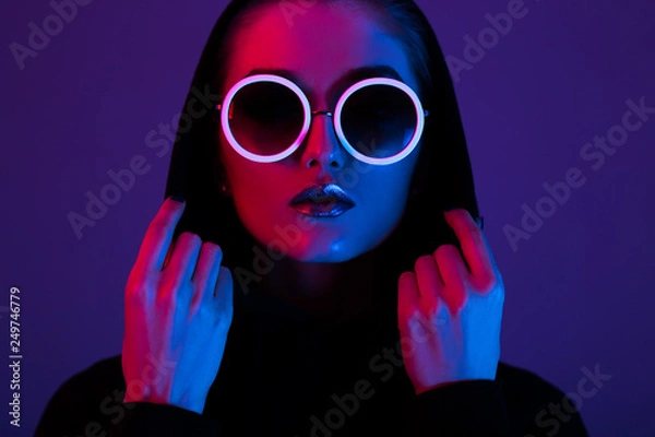 Fototapeta Portret młodej dziewczyny moda w czarnym swetrze z kapturem i okrągłymi okularami przeciwsłonecznymi w czerwonym i niebieskim świetle neonowym w studio