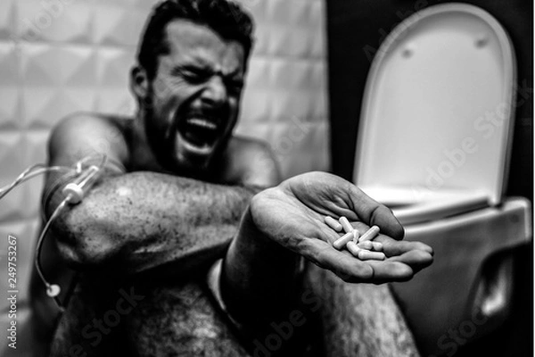 Fototapeta Balck and white picture of young man screaming and yelling on floor in rest room. He suffer. Guy show pills in hand. Drugs addiction. Hand is wrapped with plait. Another dose.