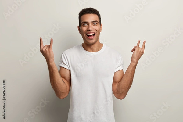 Fototapeta Happy pleased man with cheerful facial expression, makes rock n roll gesture, enjoys cool music, dressed in white t shirt, poses in studio, makes horns, has short dark hair. Body language concept