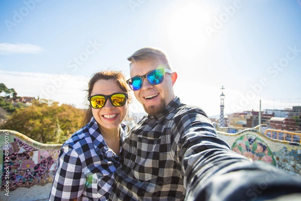 Fototapeta Funny young couple looking at camera taking photo with smart phone smiling in Park Guell, Barcelona, Spain.
