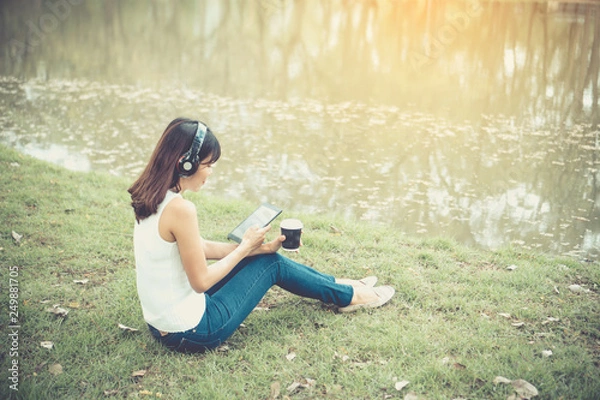 Fototapeta Beautiful girl is relaxing by listening to music. In the park there is a beautiful lake.She wears a white shirt And in her hands are choosing the song on the tablet. And she looks very happy.
