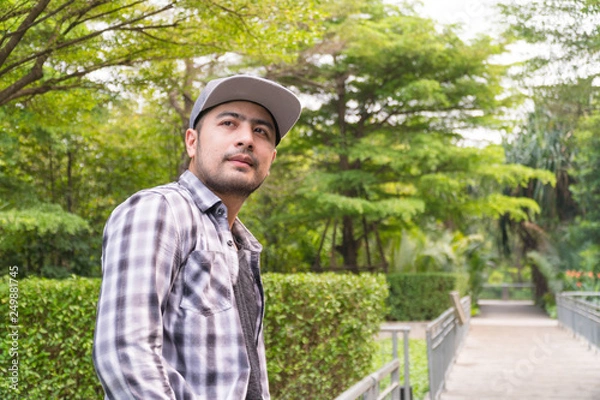 Fototapeta Portrait Handsome man : Good looking young man. He has a beard and wears a hat. He has an interesting smile. Looking for someone. Behind him, full of green trees. Copy space.