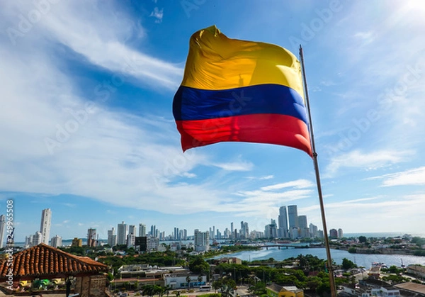 Fototapeta The Colombian flag waving in the wind. Blue sky with white clouds in the background. The city of Cartagena is in the far back. Shot in Cartagena, Colombia, USA.