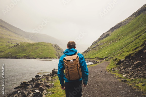 Fototapeta Young Man with a backpack from behind hiking on a trail through fjord in the wild country