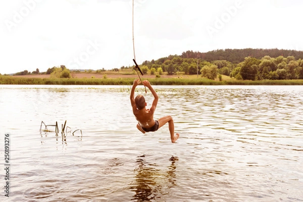 Fototapeta Teenage boy jumping in the river from the swinging rope on sunny summer day.