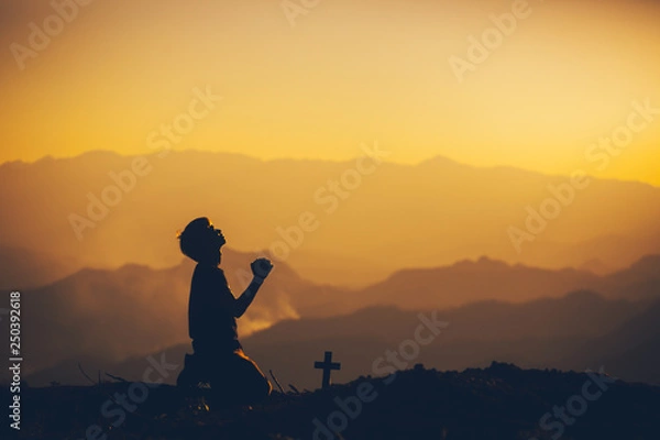 Fototapeta Young man kneeling down and praying with christian cross at sunset background. christian silhouette concept.