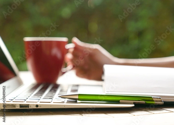 Fototapeta Work concept: hand on red mug with laptop, note pad in view and focus on pen
