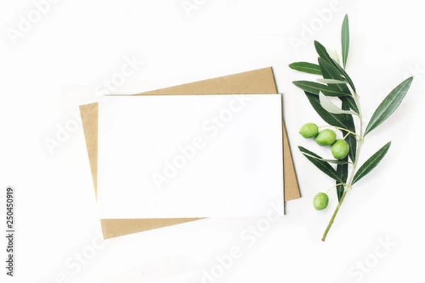 Fototapeta Feminine stationery, desktop mock-up scene. Blank horizontal greeting card and craft envelope with olive branch.White table background. Flat lay, top view.