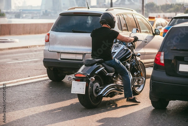 Fototapeta rear view of a handsome biker dressed in a black T-shirt and black helmet jeans and sneakers. He is riding on cruiser motorcycle on background of 2 cars. Traffic stop