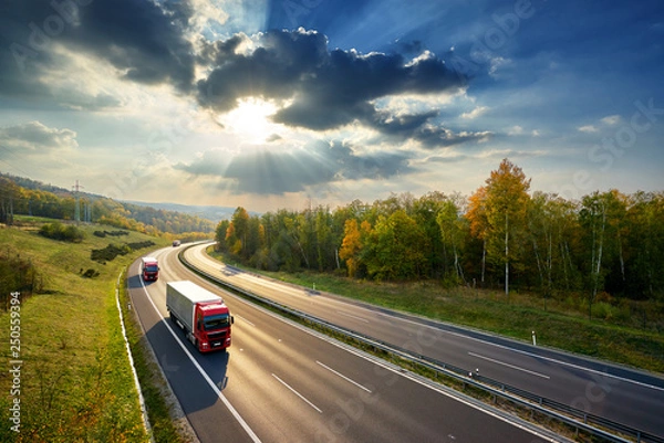 Fototapeta Trzy czerwone ciężarówki jeżdżące po asfaltowej autostradzie pomiędzy lasem liściastym w jesiennych barwach w promieniach zachodzącego słońca i dramatycznych chmur. Widok z góry.