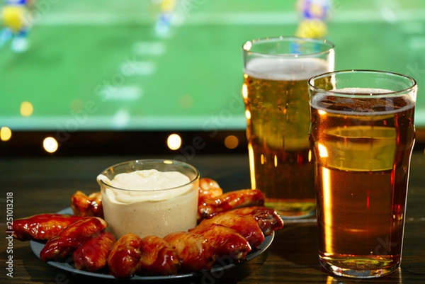 Fototapeta Hot barbecue chicken wings with 2 beer glasses on a dark wooden table served with honey mustard sauce. Football on a background, high resolution