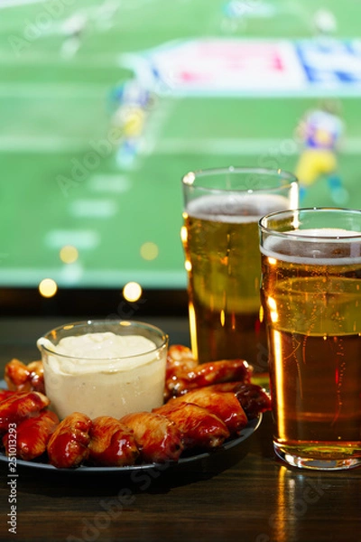 Fototapeta Hot barbecue chicken wings with 2 beer glasses on a dark wooden table served with honey mustard sauce. Football on a background, high resolution