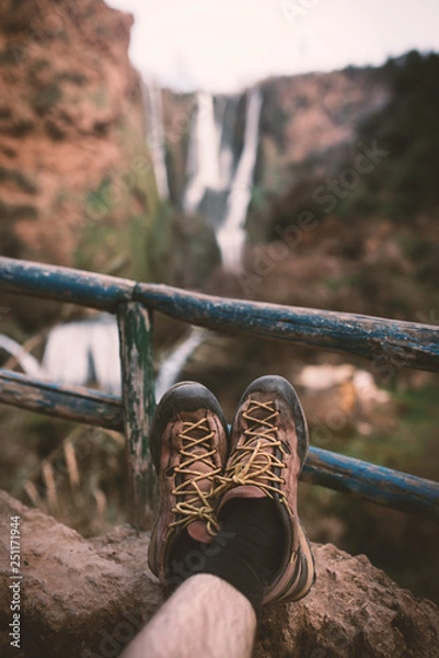 Fototapeta Man´s trekking shoes in a nature of Morocco - Ouzod falls. Close up of hiking boots against waterfall. Man hiking enjoying a view after trek.  