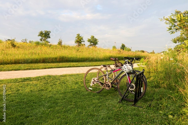 Fototapeta Beautiful rural summertime evening nature background. Scenic view with wild grasses field, rural road and pair of parked bicycles in a foreground during sunset. Healthy lifestyle concept.