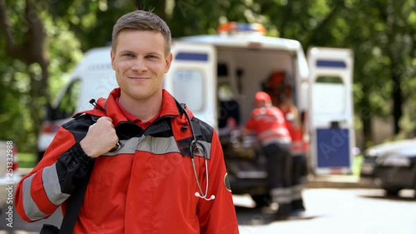 Fototapeta Male doctor smiling into camera, ambulance crew working, blurred on background