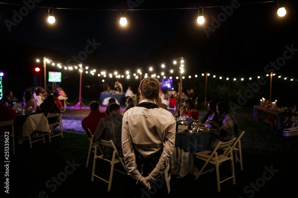 Obraz Waitress is standing at the foreground. A lot of tables with many guests sitting behind them are at the background.