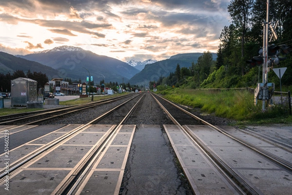 Fototapeta Parallel Railroad Tracks through a Town Centre with Towering Mountains in Background at Dusk. Revelstoke, BC, Canada.