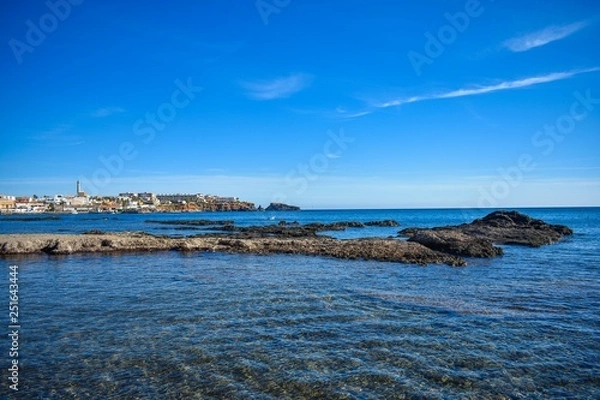 Fototapeta Crystal clear water envelop dark grey rocks that just rise above the surface, with the small village of Cabo de Palos, Spain in the background.