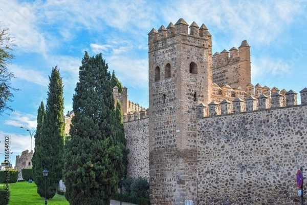 Fototapeta The defensive stone walls of Toledo, Spain, showing the battlements on top and a guard tower with the green lawn that now surrounds the city.