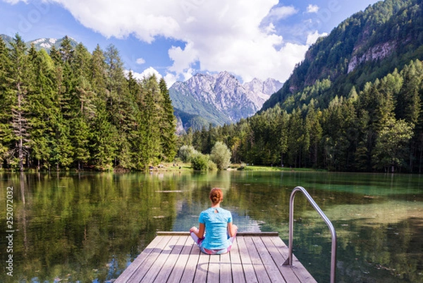 Fototapeta Young woman is sitting on a pier over the lake looking at the magnificent view  in Jezersko, Slovenia. Woman relaxing near the lake looking at the view. Travel adventure and freedom concept.