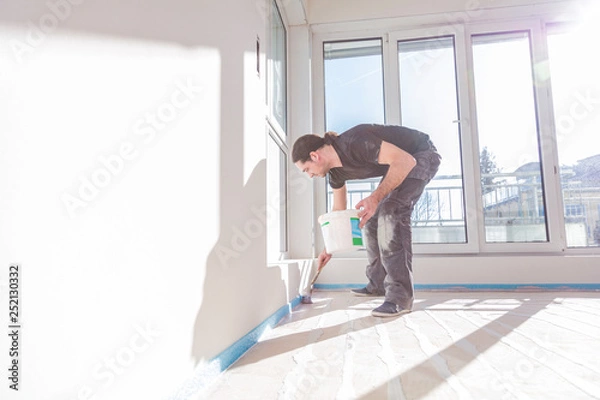 Fototapeta House under construction. Worker puts primer with roller on concrete floor.
