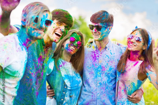 Fototapeta Cheerful and happy friends soiled by bright colorful paints making selfie and laugh on spring summer festival on nature. Company of young people having fun with holi paints. Holi party concept.