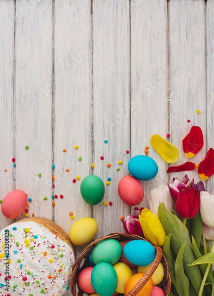 Fototapeta Colorful easter eggs,cake,spring tulips  on wooden texture background.On a white wood table,colored eggs,flowers,bread.Happy religious day,traditional for people. Top view.Copy space.