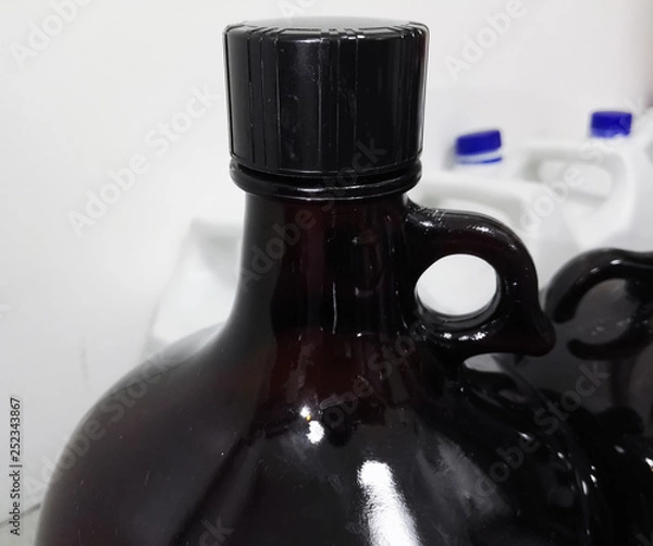Fototapeta Large amber bottle (liter) to contain chemical reagents in a scientific laboratory or for disposal of hazardous waste, with a background of other white containers with a blue cap