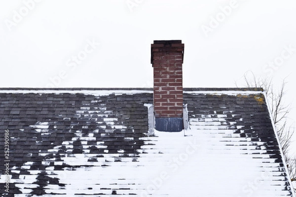 Obraz Brown brick chimney on grey asphalt shingles roof partly covered with snow in winter against sky with copy space for text.