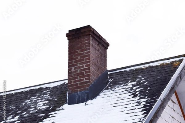 Fototapeta Brown brick chimney on grey asphalt shingles roof partly covered with snow in winter against sky with copy space for text.