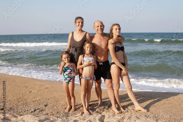 Fototapeta Group portrait of a healthy family of 5 people, mom, dad and three daughters on a sunny beach are standing on the sand in bathing suits, and smiling with happy faces