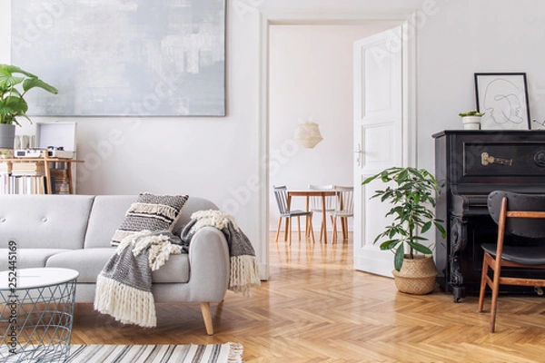 Fototapeta Modern scandianvian decor of living room with design sofa with elegant blanket, coffee table and bookstand on the white wall. Brown wooden parquet. Concept of minimalistic interior with piano. Mock up