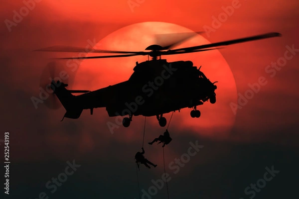 Obraz Shot of rescue helicopter hopping in front of the big sun with two soldiers rappelling from high
