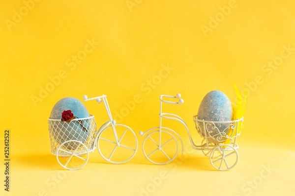 Obraz Two Easter eggs ride towards each other on bicycles on a yellow background.