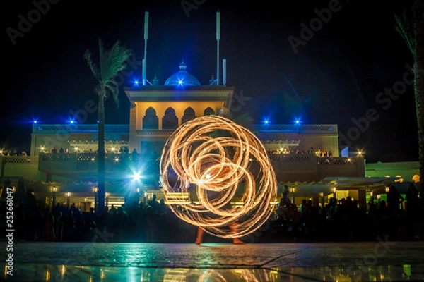 Fototapeta Night performance fire show in front of a crowd of people on the street in Egypt, palace and palm trees in the soft focus background.