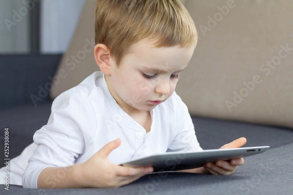 Fototapeta Little blonde boy using touchscreen tablet at home. Happy smart child watching tutorials or playing game on tablet computer. Education, technology, children's security and fun concept.
