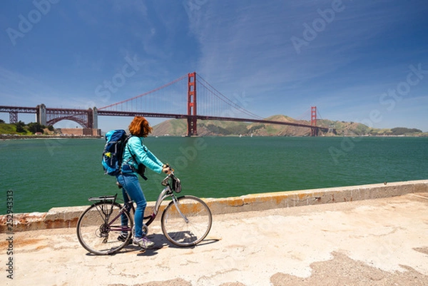 Fototapeta Young woman is sitting on a bicycle looking at the Golden Gate bridge from a pier in San Francisco, United States of America