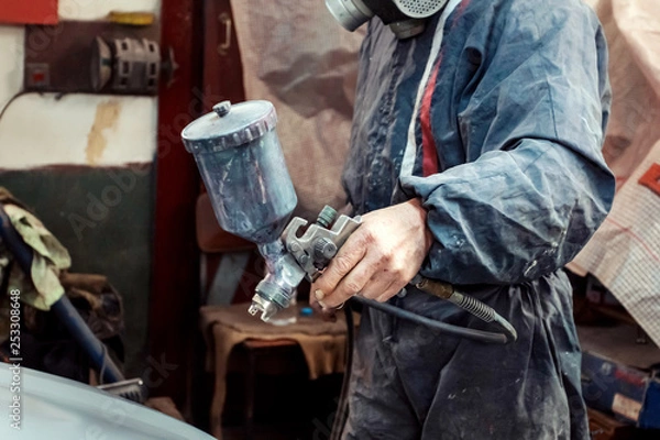 Obraz Man with protective clothes and mask painting car using spray compressor. Selective focus on hand.