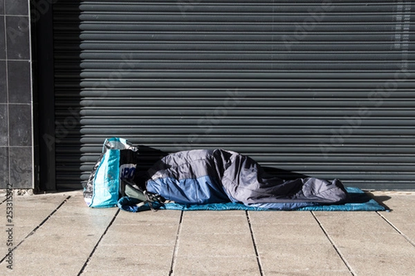 Fototapeta Homeless person asleep in a sleeping bag on a pavement sidewalk in front of a metal shutter.  Face not visable.