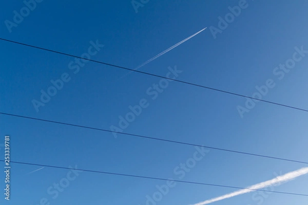 Fototapeta Airplanes in a blue sky with white contrails behind them cross electric wires. Norilsk