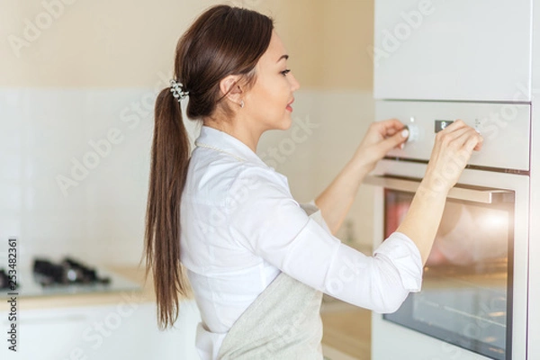 Fototapeta cheerful Asian girl turning on the oven in the kitchen. close up side view photo. chef has put the dish in the oven to preheat it