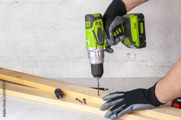 Fototapeta The builder holds in his hand an electric screwdriver on the background of a concrete wall. Screws screw into a wooden beam. DIY