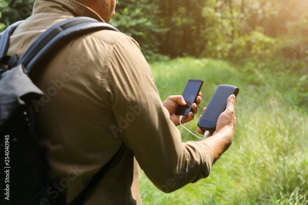 Fototapeta Tourist with smartphone and battery charger is looking for the way in summer forest. Guy with powerbank is lost in the nature. Modern technologies concept to stay always online and in touch.